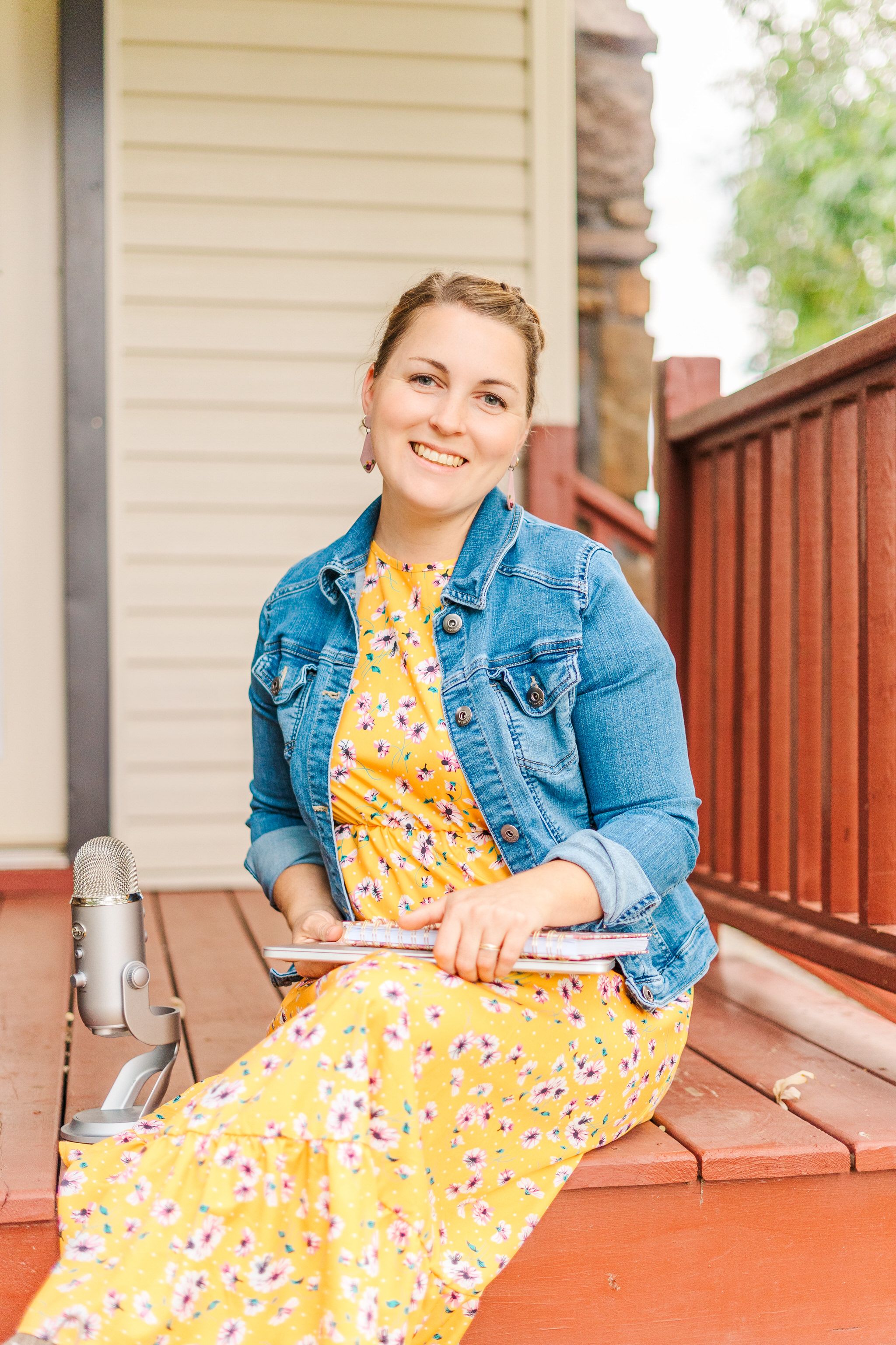 Someone sits on wooden deck steps wearing a yellow floral dress and denim jacket on a sunny day.