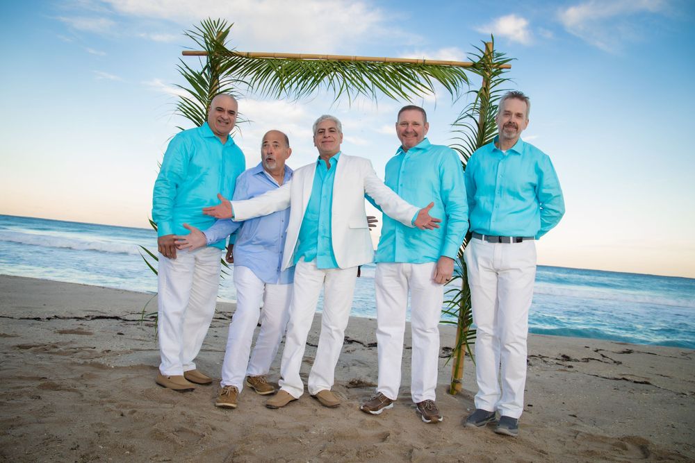 A group in coordinated turquoise and white outfits poses together on a sunny beach with palm trees and ocean views.