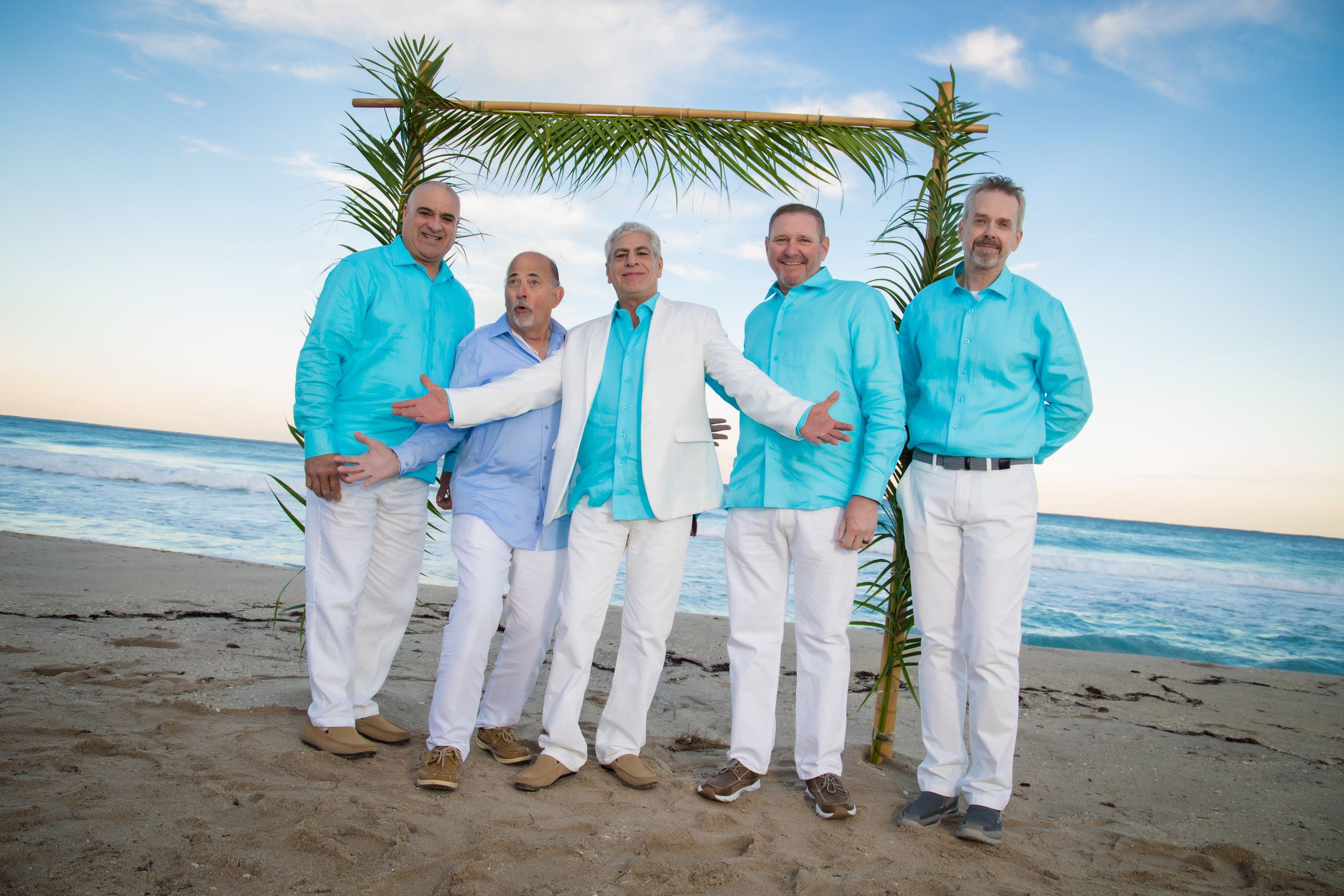 A group in coordinated turquoise and white outfits poses together on a sunny beach with palm trees and ocean views.