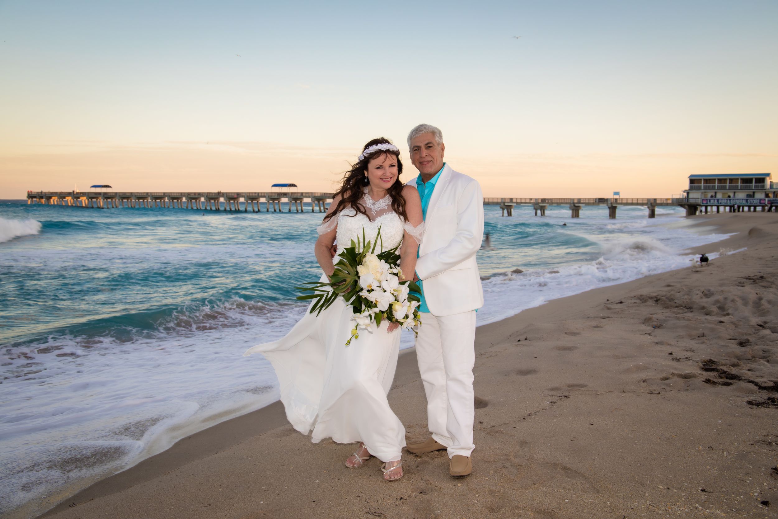 A couple in white attire walks along a sandy beach during sunset with waves crashing in the background.