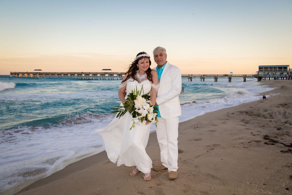A couple in white attire walks along a sandy beach during sunset with waves crashing in the background.