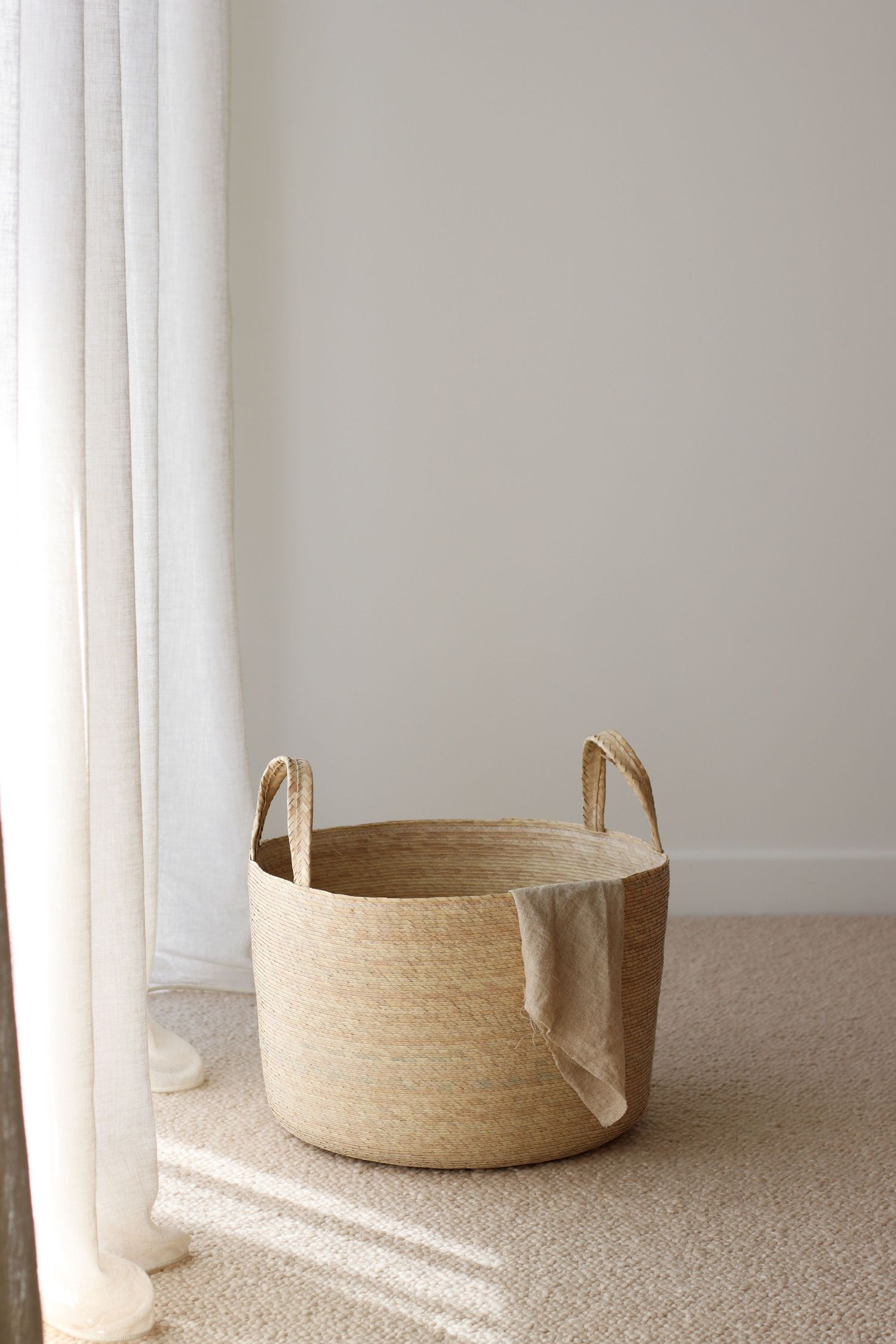 Natural woven basket with handles sits near white curtains in minimalist setting.