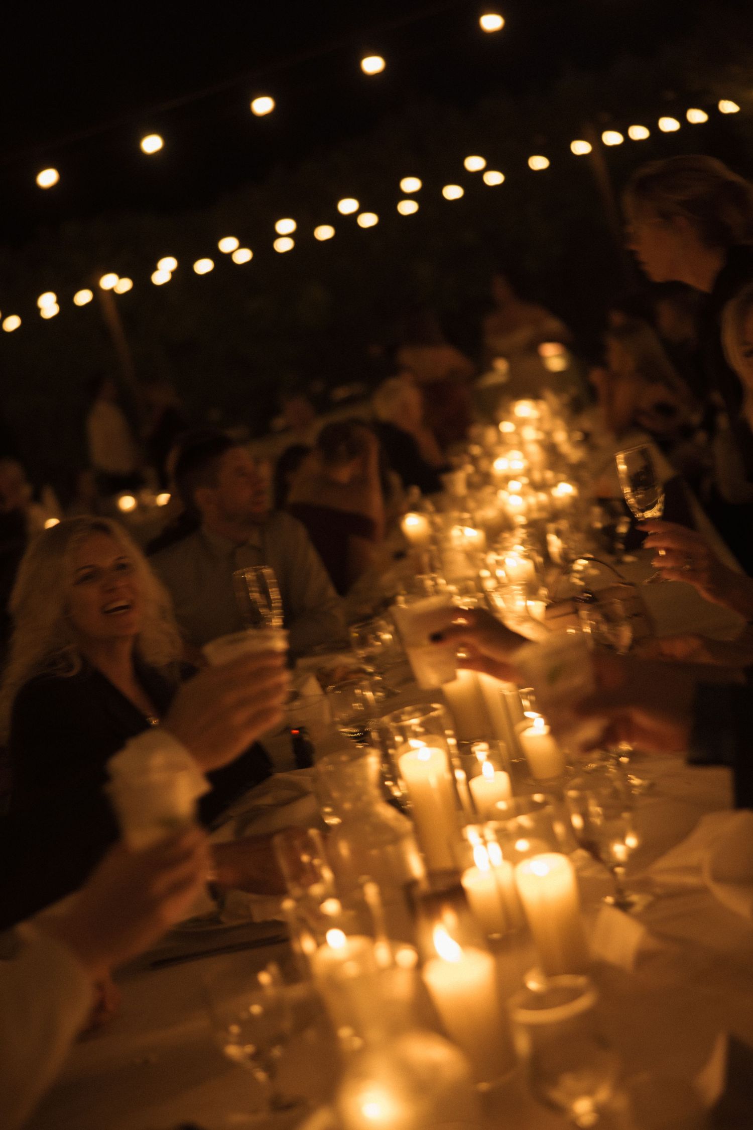 Candlelit wedding reception with guests cheering at a long table, warm twinkle lights and romantic motion blur