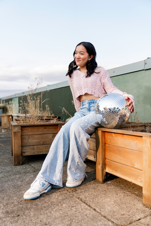 High school senior posing on a rooftop with a disco ball, rocking a retro vibe in wide-leg jeans and a pink sweater – stylish senior photos.