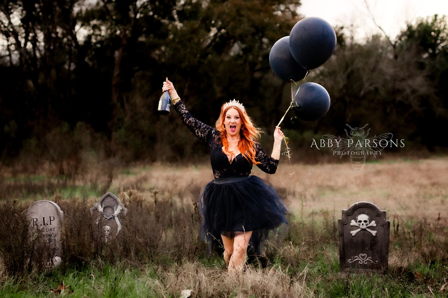 A figure in a black tulle dress celebrates in a spooky graveyard setting with navy balloons and tombstones at dusk.