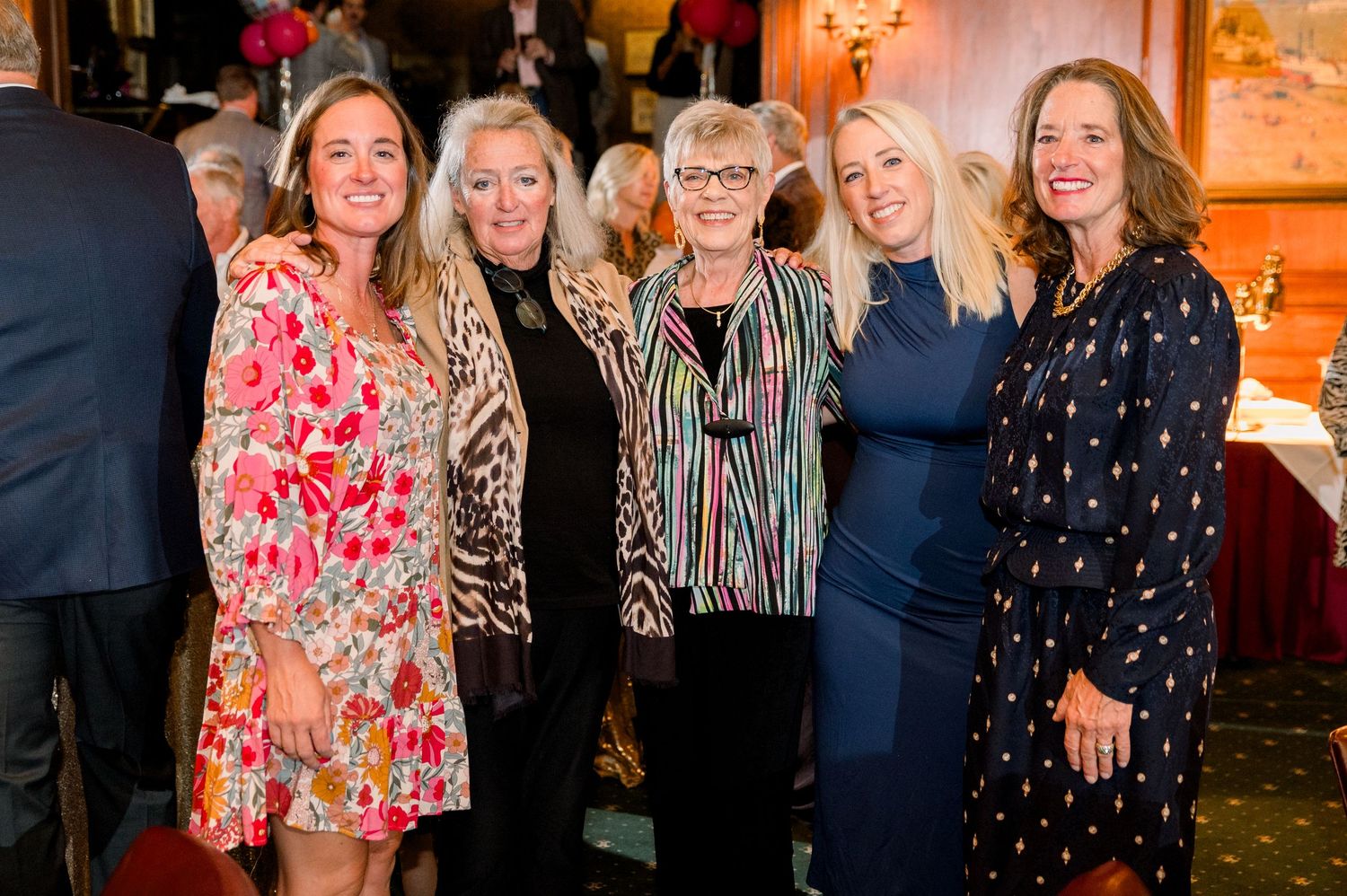 Five women smiling together at an elegant indoor event, dressed in colorful and stylish outfits.