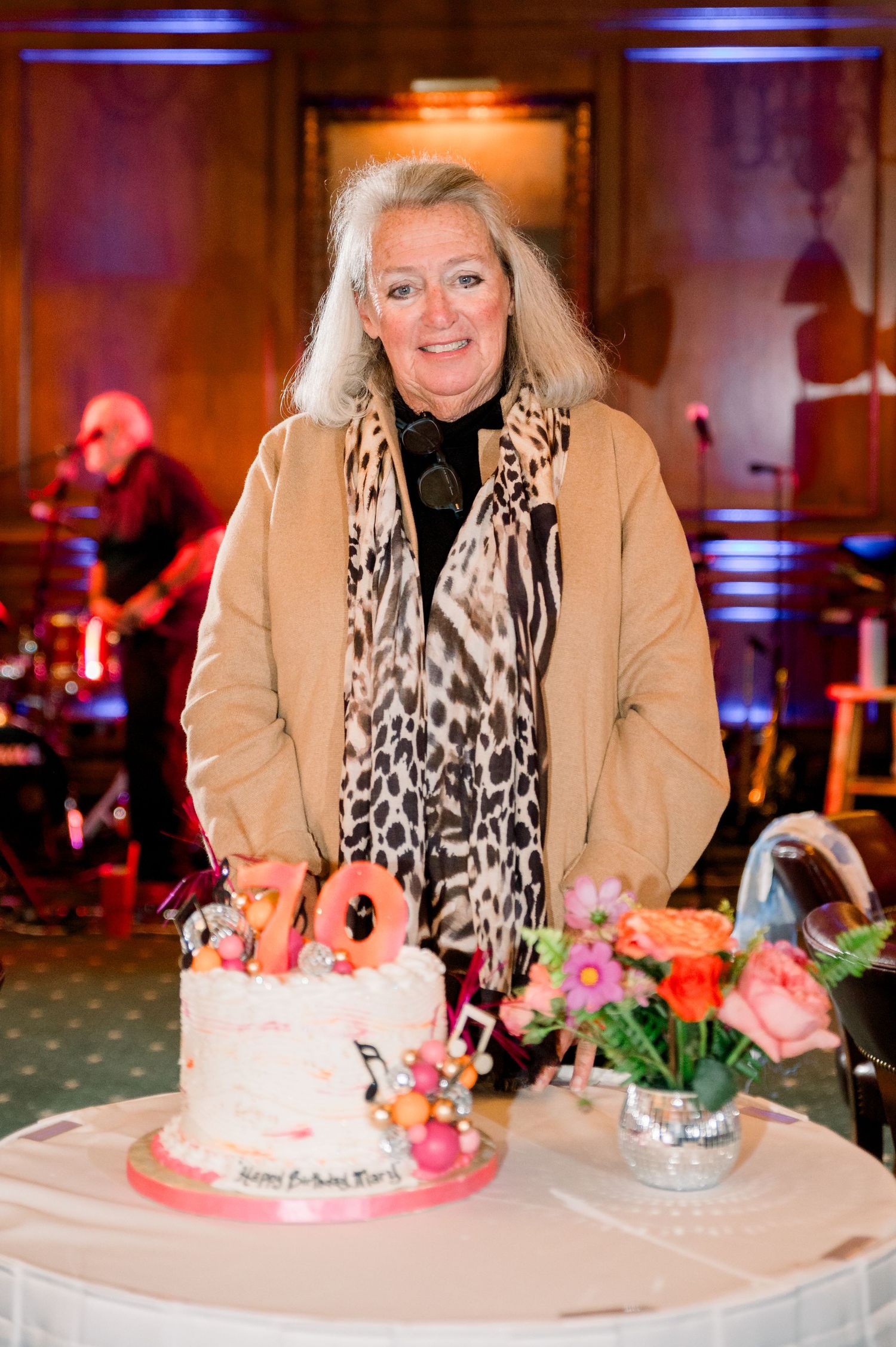 Woman smiling behind a 70th birthday cake at an elegant party venue with a live band in the background.