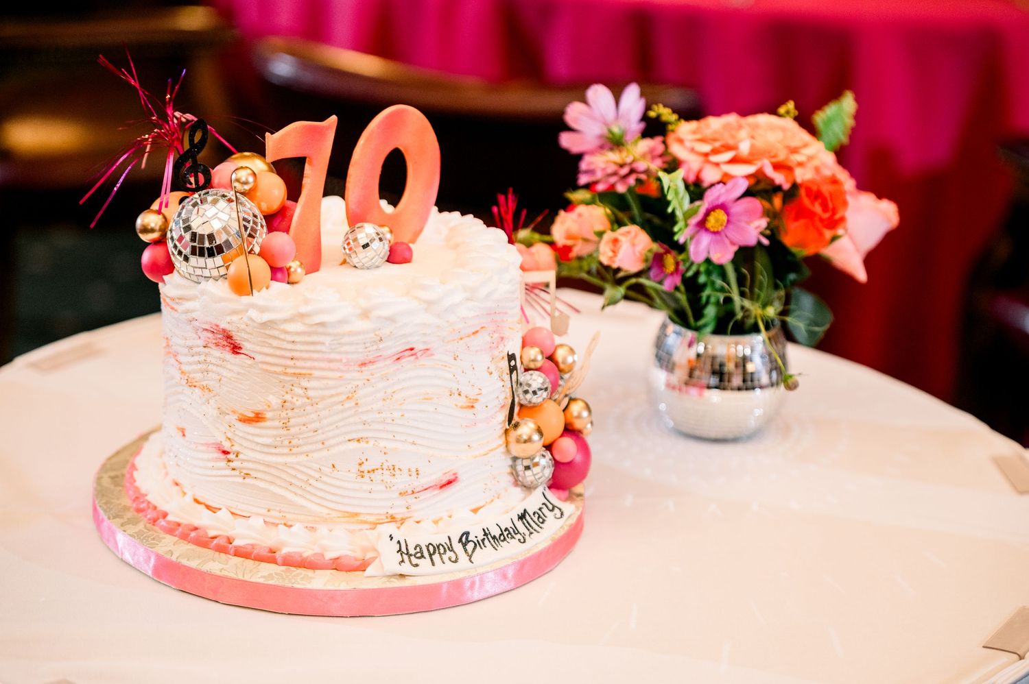 Elegant 70th birthday cake with pink number candles, disco ball decorations, and floral arrangement on white table.