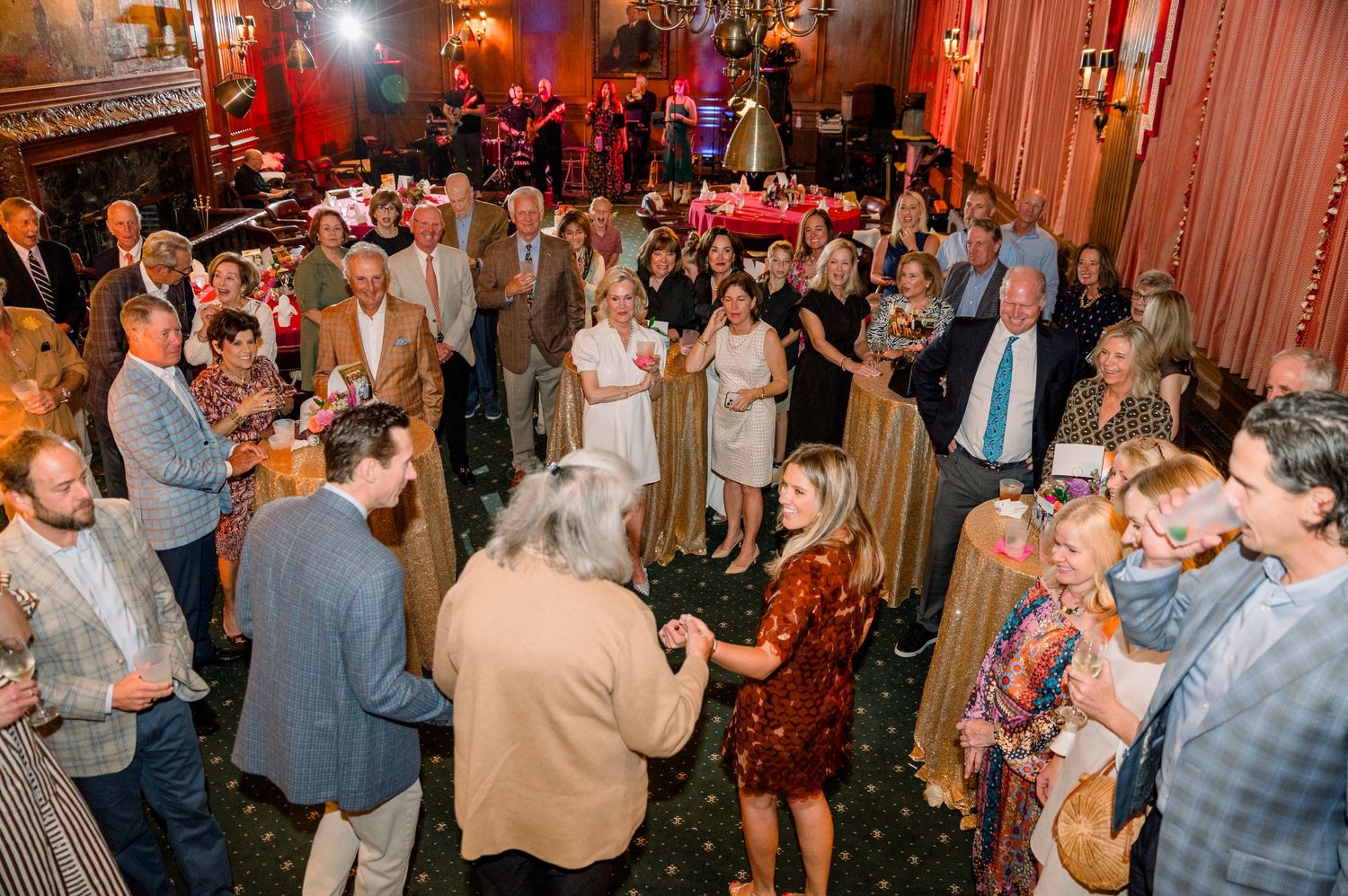 Elegant indoor party with guests mingling and dancing in a grand decorated ballroom with red lighting.