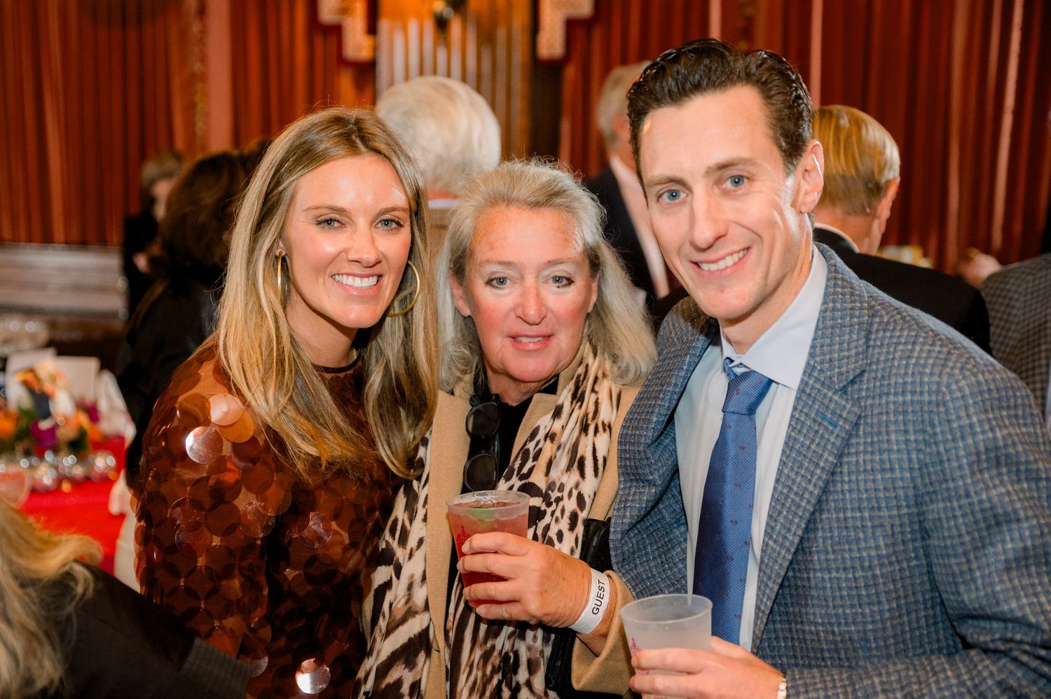 Three smiling guests pose together at an elegant indoor social event, holding drinks in a warmly lit venue.