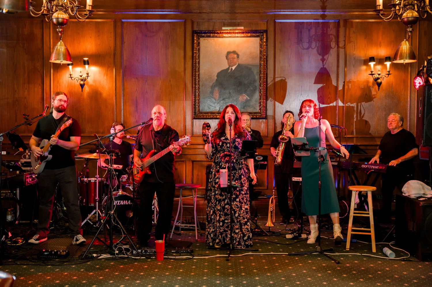 Live band performing on stage in an elegant wood-paneled room with portrait painting, featuring vocalists and musicians.