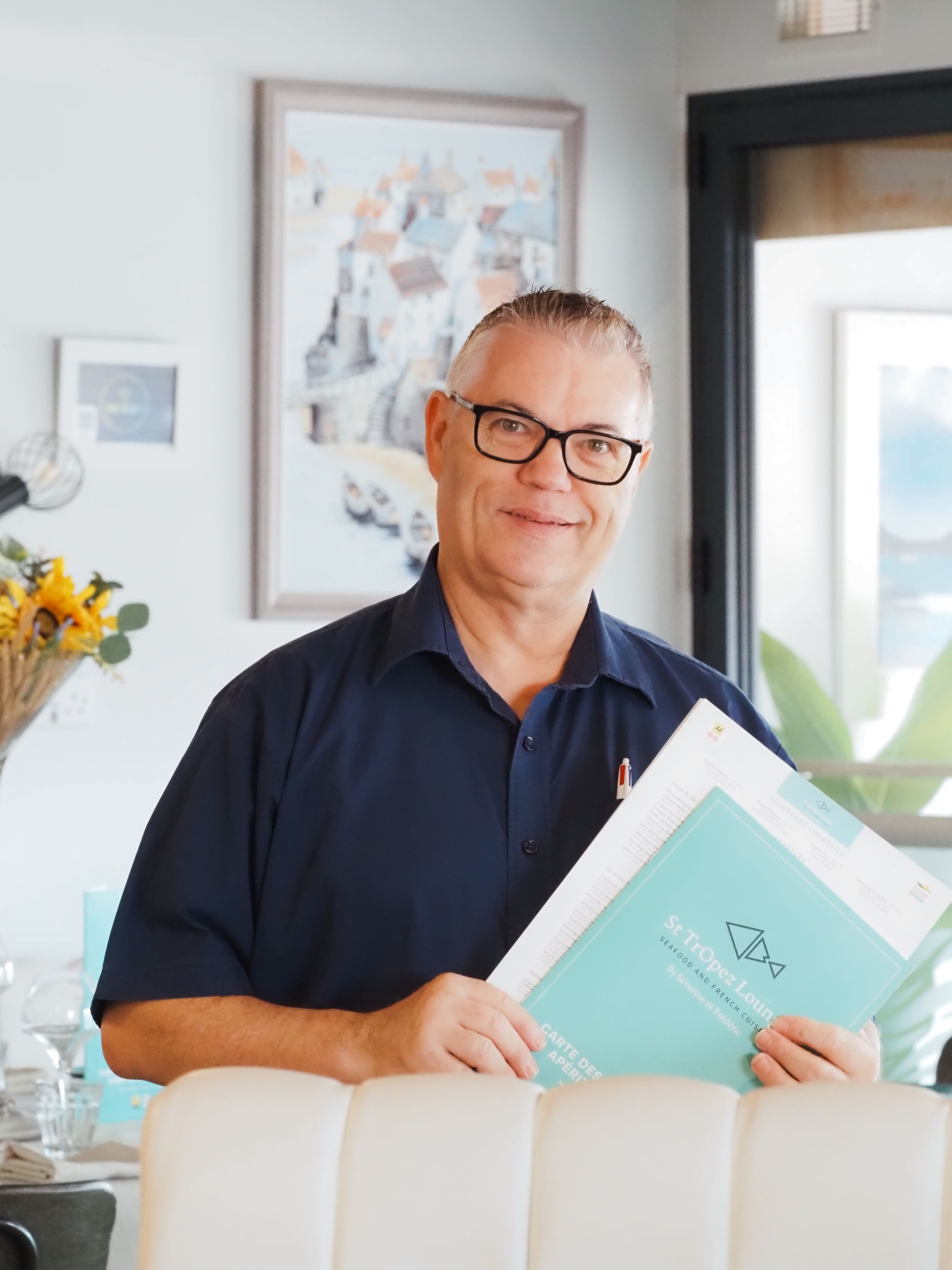 A person wearing glasses and navy shirt reads a turquoise book in bright interior with windows and wall decor.