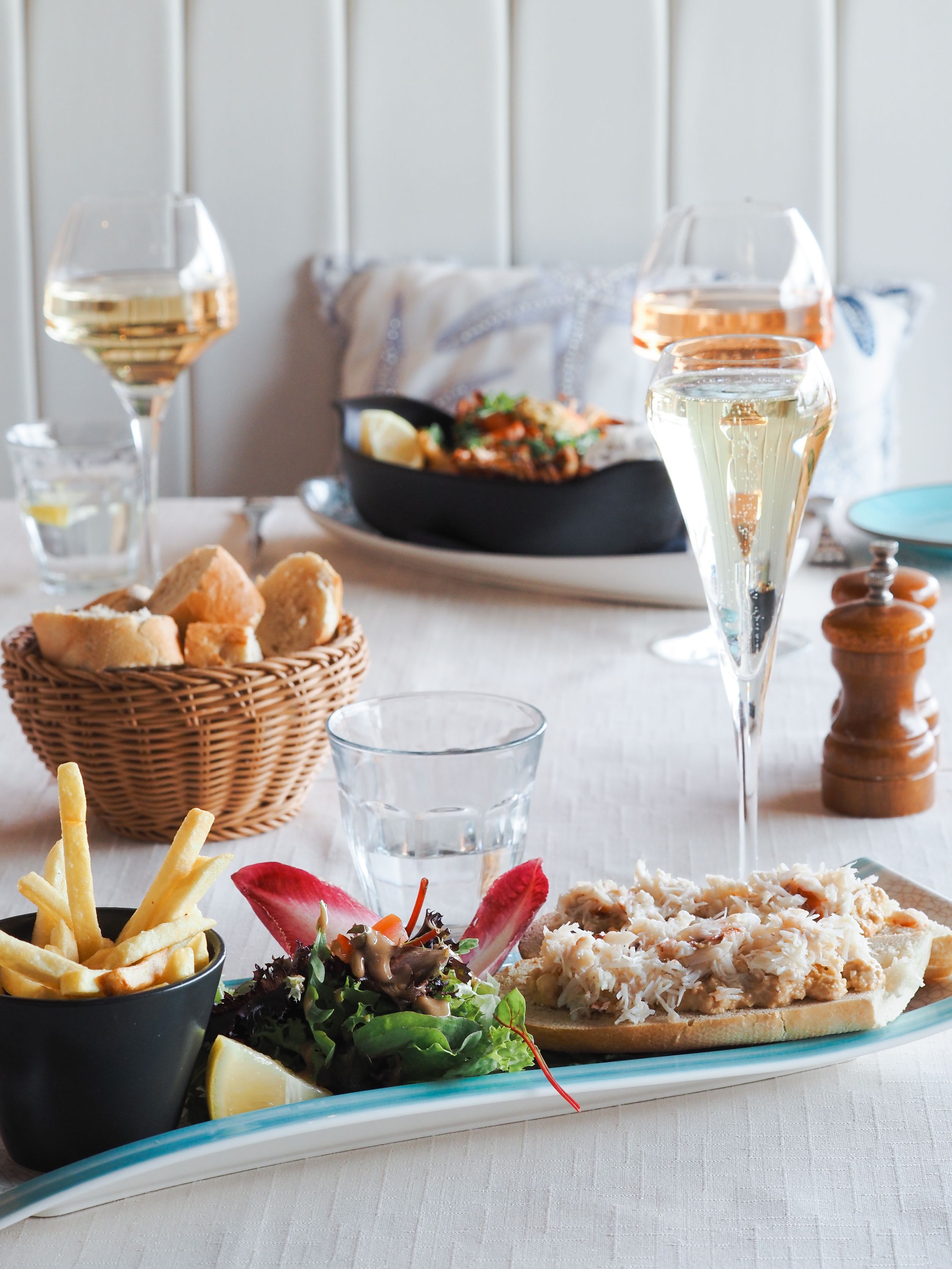 Array of appetizers and bread baskets displayed on white tablecloth with wine glasses and coastal dining presentation.