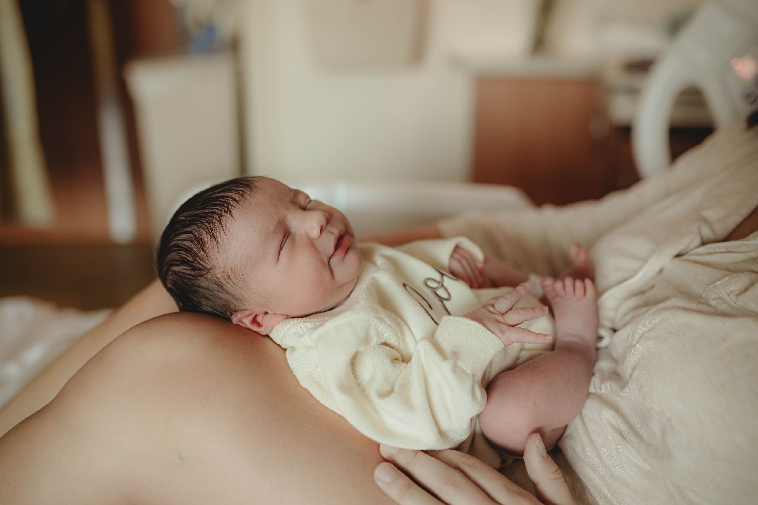 Sleeping newborn in soft cream clothing lying on white bedding in natural light.