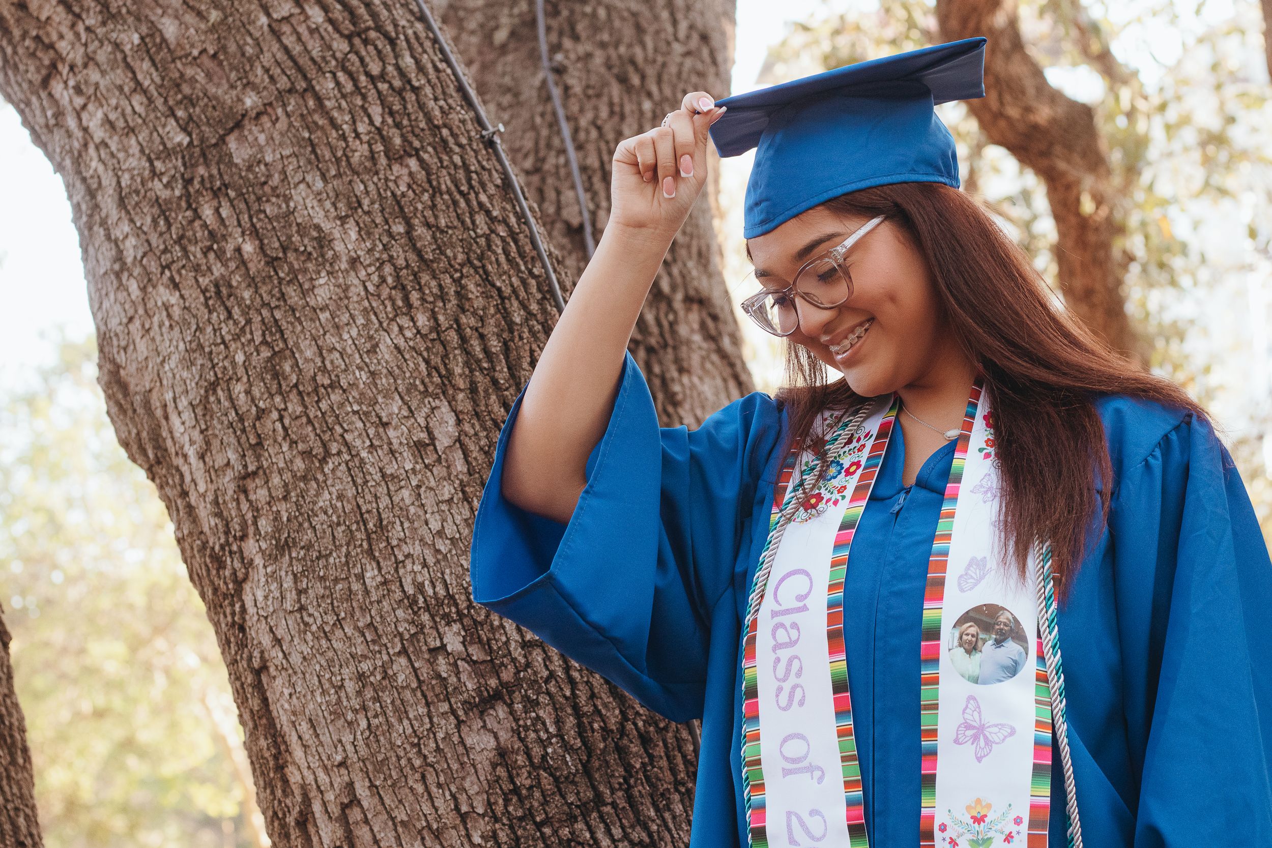 Smiling graduate in blue cap and gown leaning against a tree during graduation photoshoot.