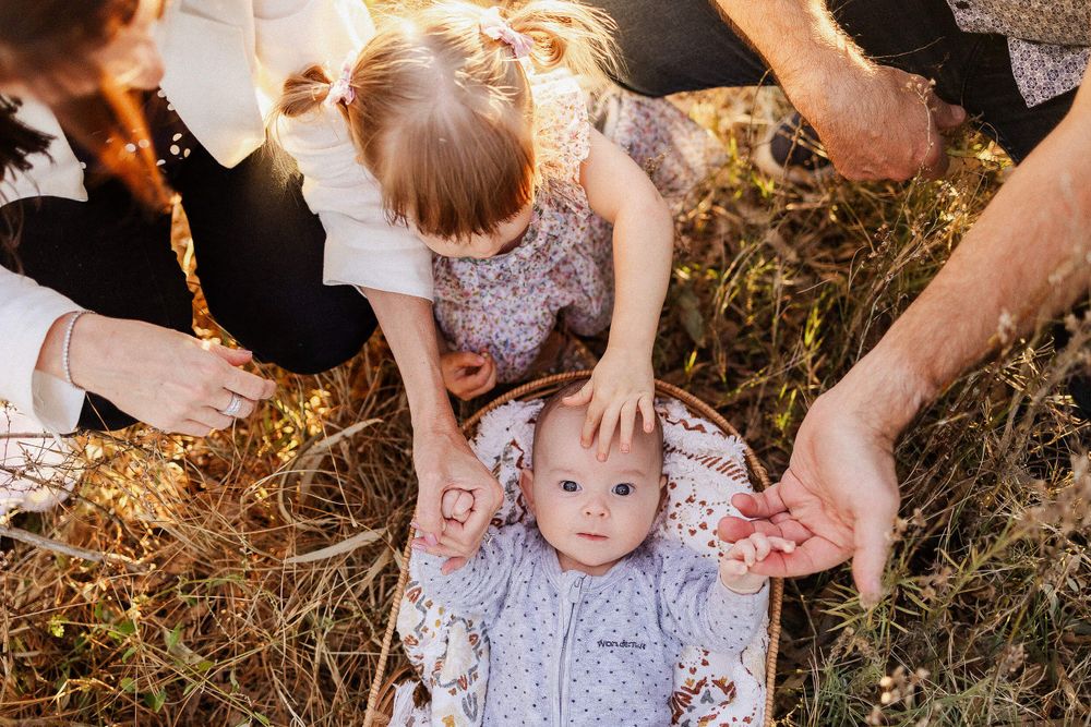 A family sits together in golden sunlight on dry grass, wearing light coloured clothes while sharing a tender moment.