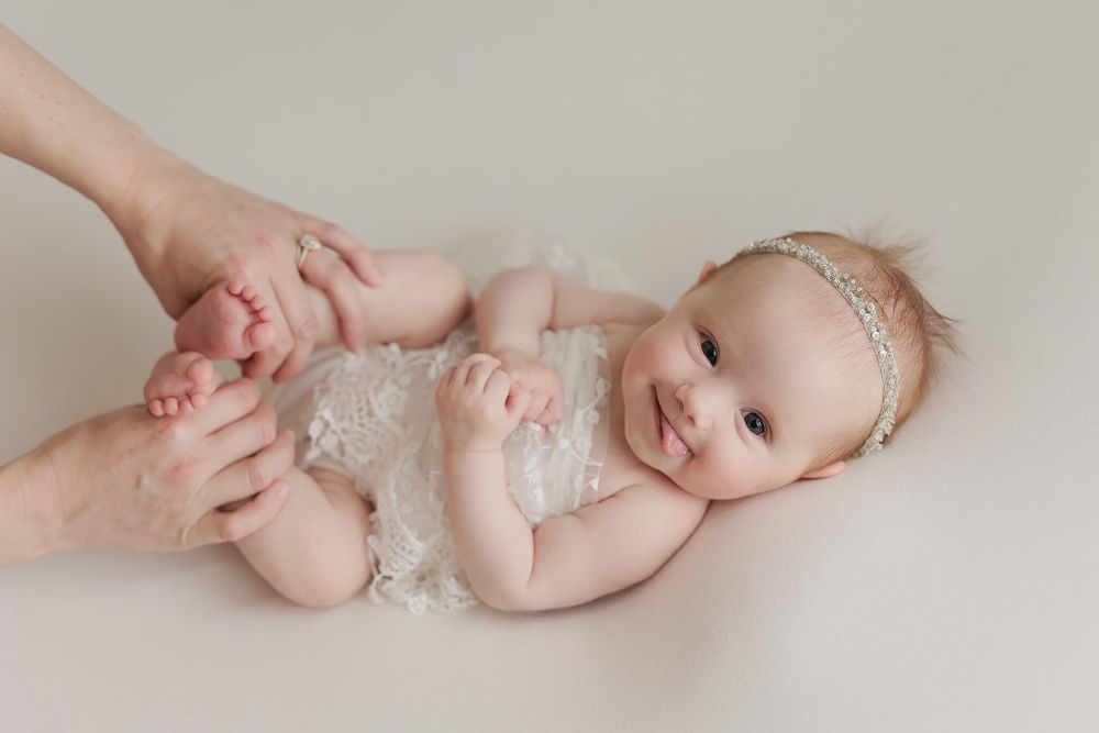 A baby in white lace romper lying on beige backdrop while gentle hands hold their feet during newborn photoshoot.