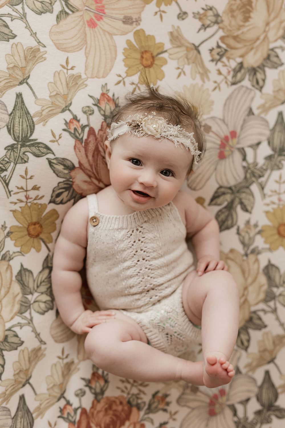 Sweet baby wearing cream knit romper and lace headband lies on vintage floral print background.
