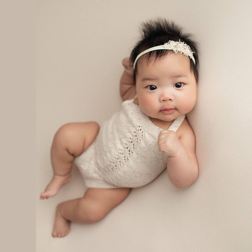 Adorable baby wearing white lace romper and floral headband poses laying on beige seamless backdrop during photo session.