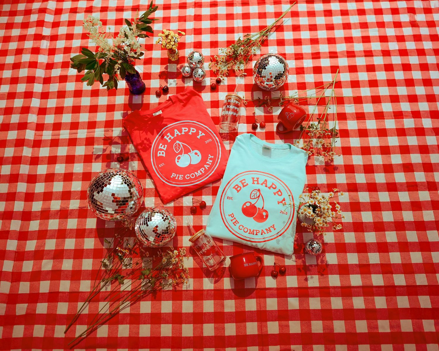 Two Be Happy t-shirts in red and mint displayed on a red gingham tablecloth with disco balls and dried florals.