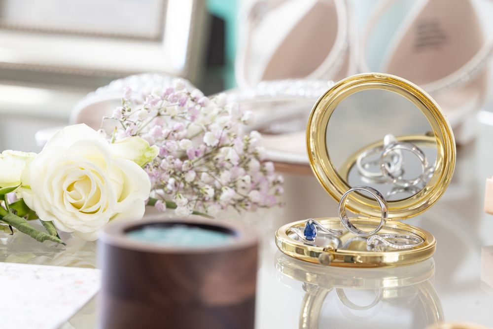 Close-up of wedding rings and delicate white flowers arranged on a reflective surface in soft natural light.