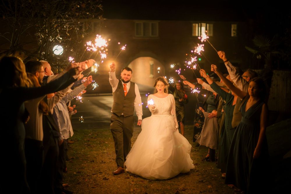Newlyweds walk between rows of wedding guests holding sparklers during an evening outdoor celebration.