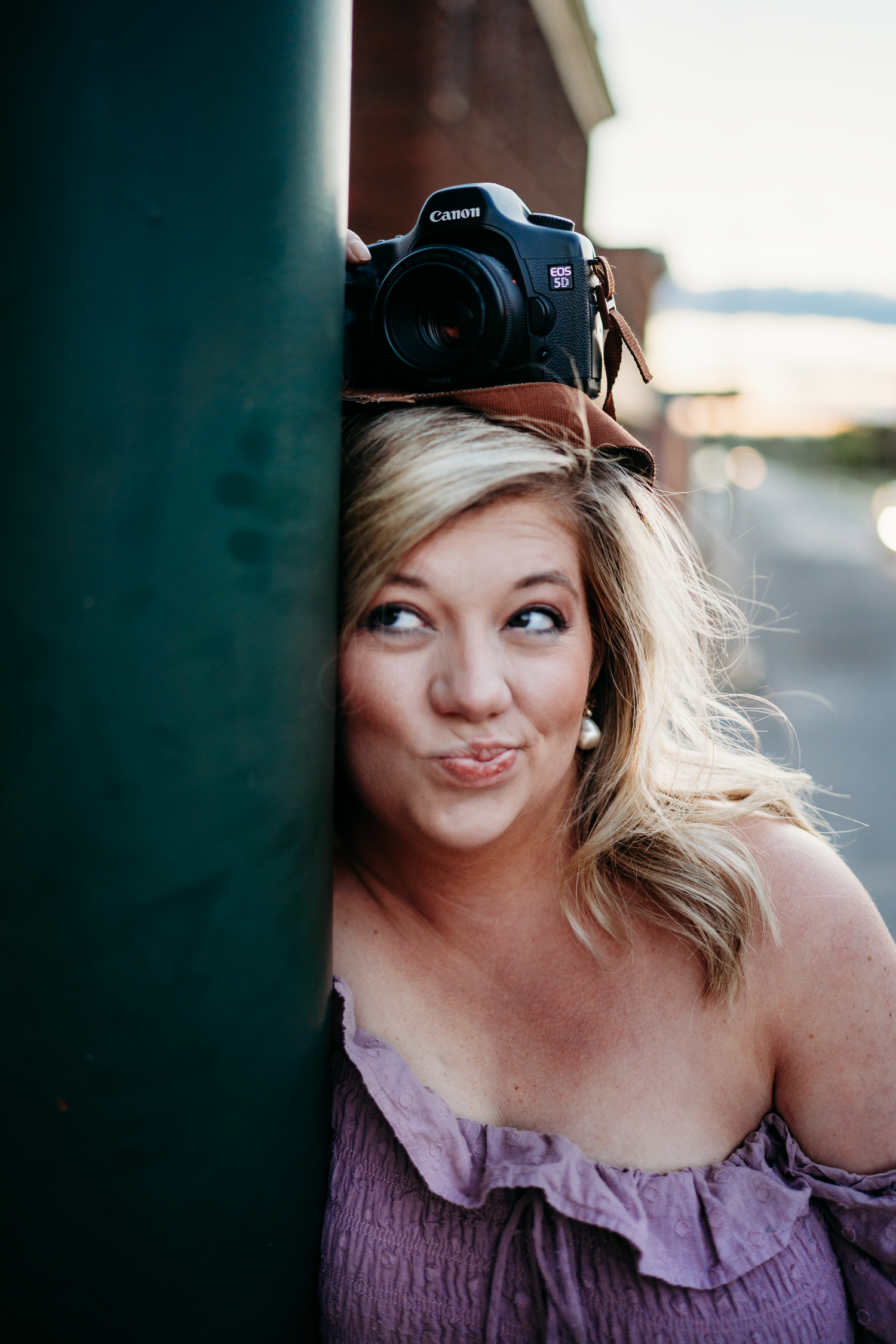 Playful blonde female photographer balancing Canon DSLR camera on head while peeking around green pole.