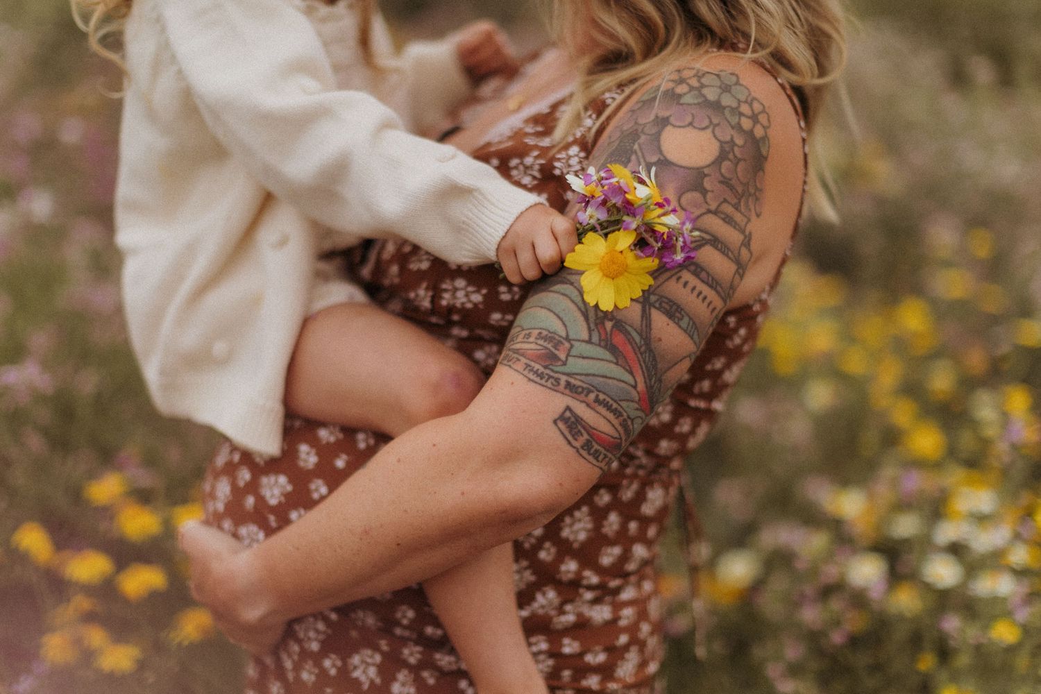 A mother and child in brown matching dresses sit among yellow wildflowers in a meadow during golden hour.