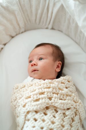 Newborn baby wrapped in a white knit blanket lying on soft bedding.