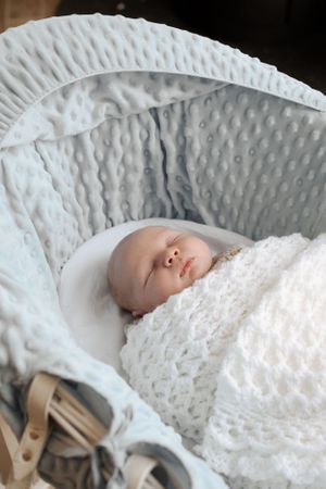 A sleeping infant wrapped in white lace blankets in a textured bassinet.