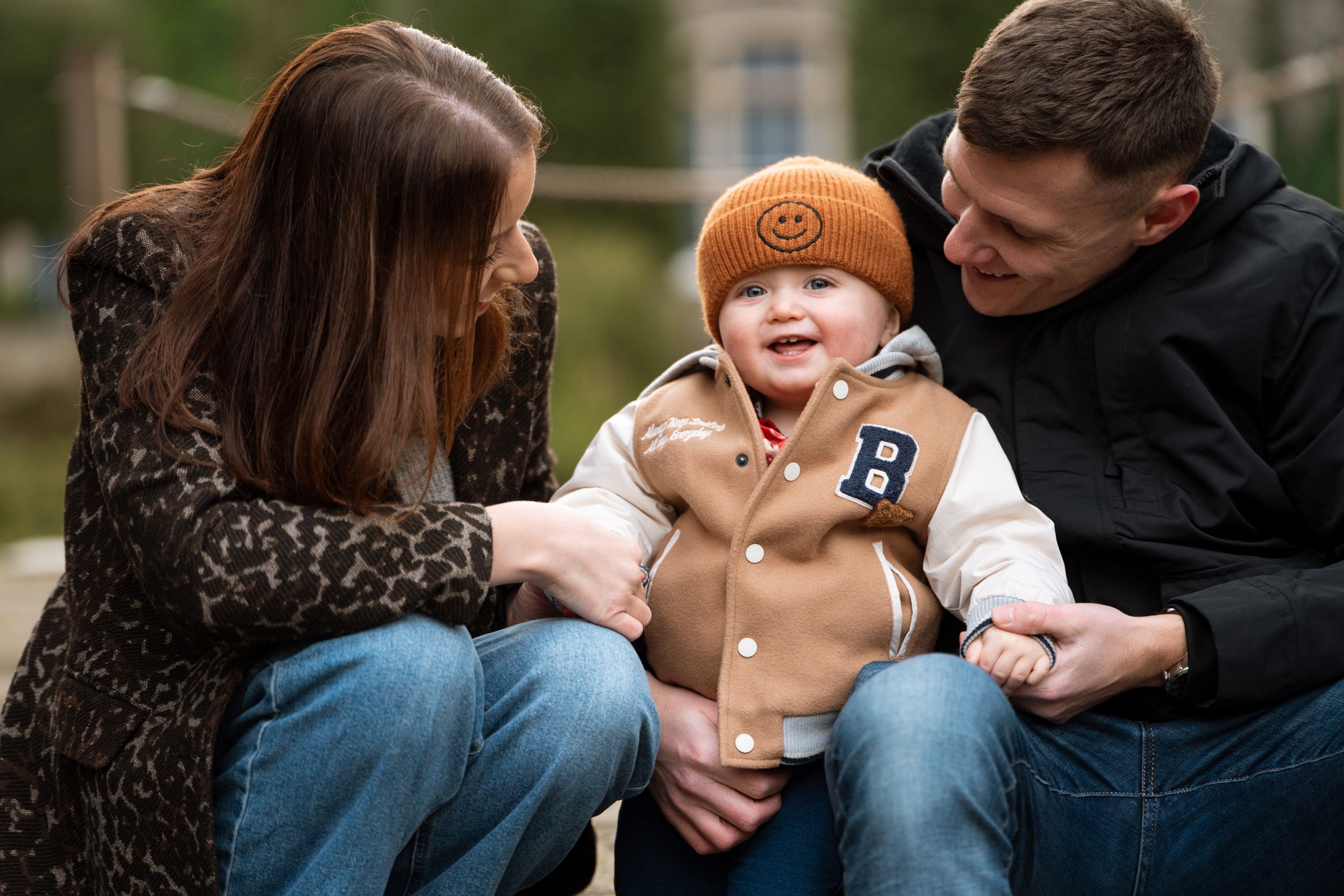 Baby wearing orange beanie and tan coat playing outdoors with parents.