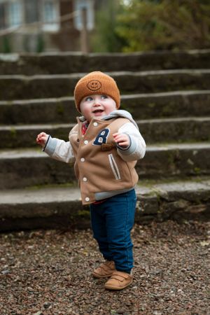 Smiling baby in brown coat and orange beanie standing on gravel path.