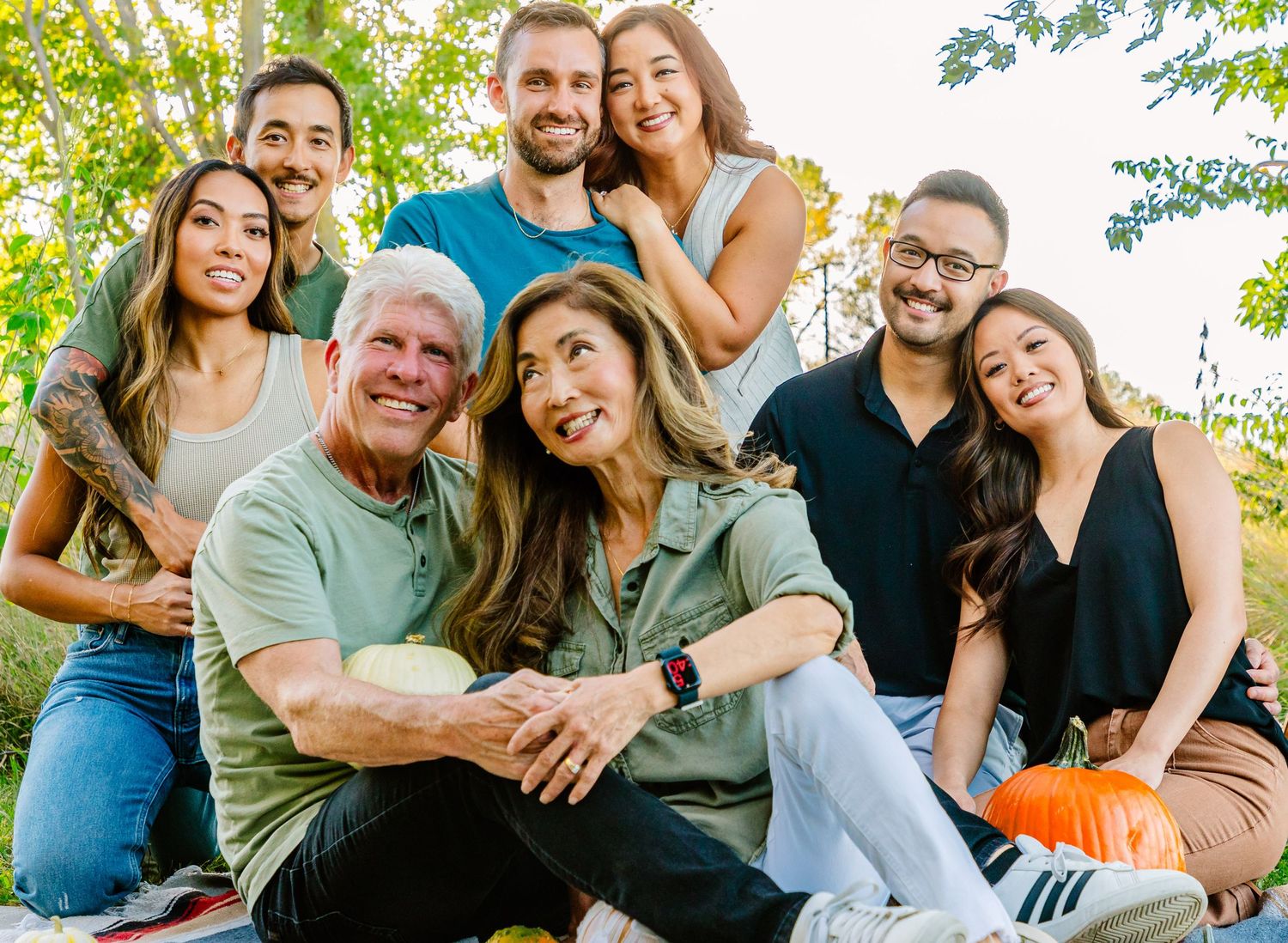 A group of people smiling and posing together outdoors on a sunny day with green foliage in the background.