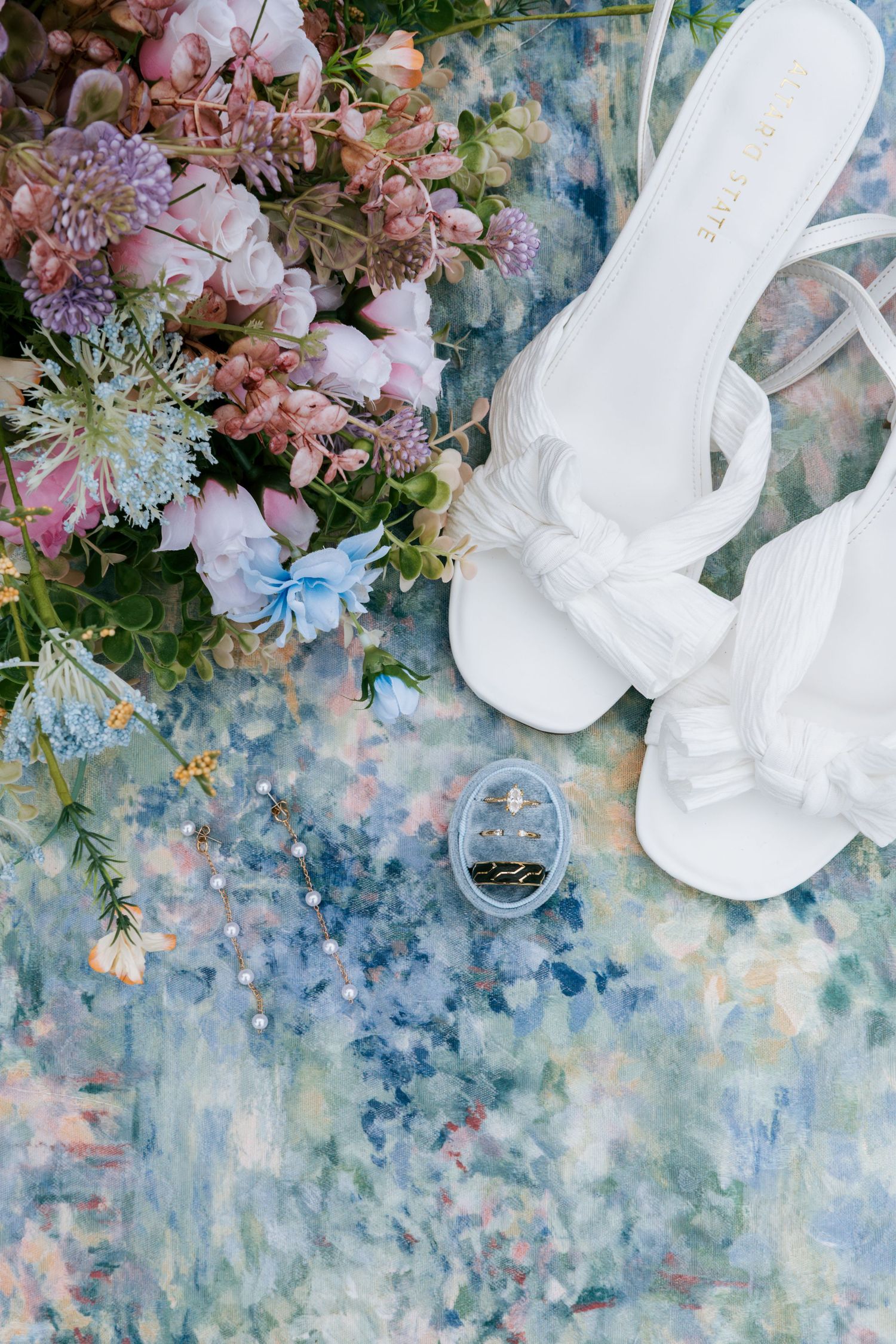 Flat lay of white bridal sandals, pink and blue wildflower bouquet, and diamond ring on blue backdrop Greensboro NC