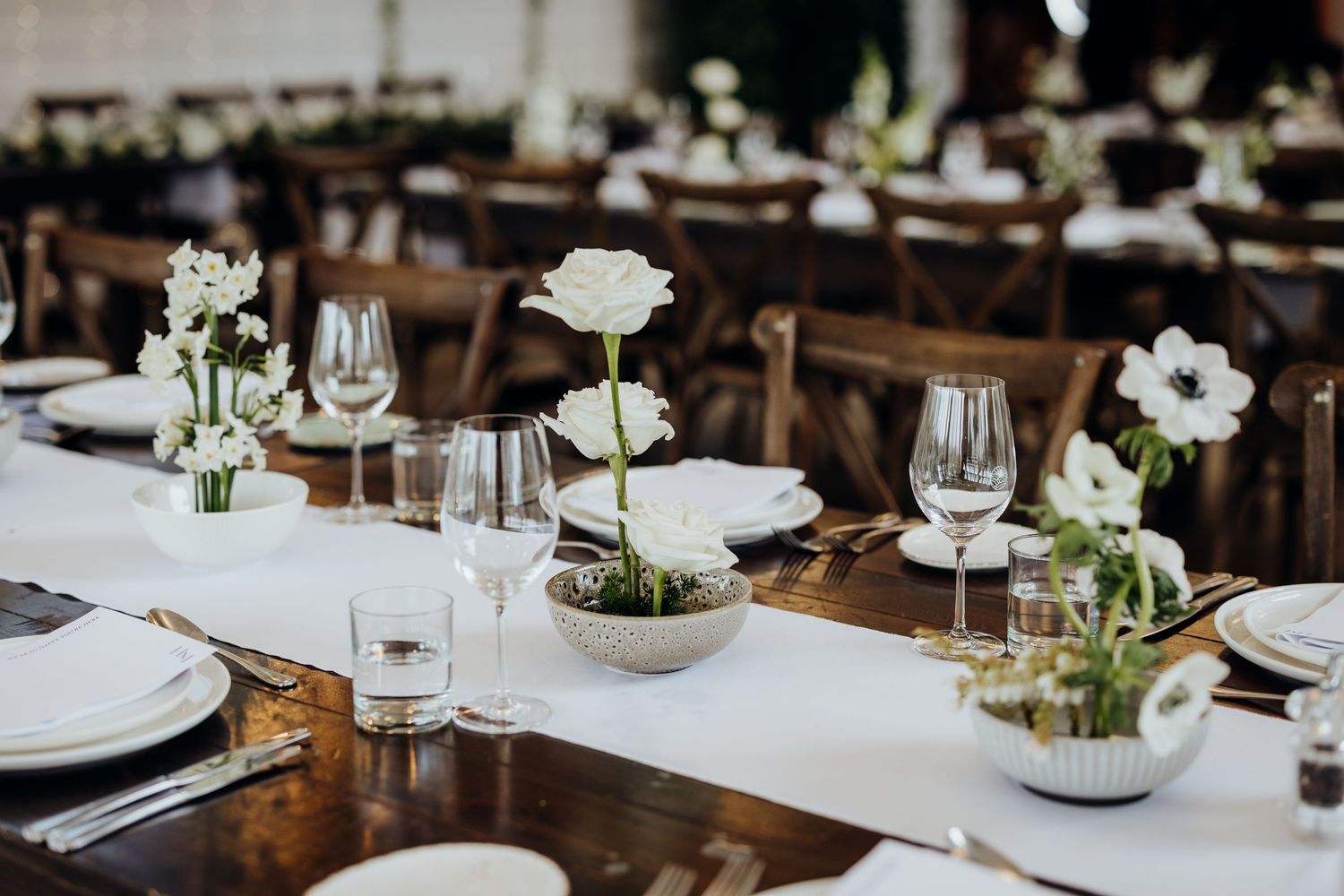 Reception table settings with white floral centerpieces, rustic wood tables, and elegant glassware at a wedding venue.