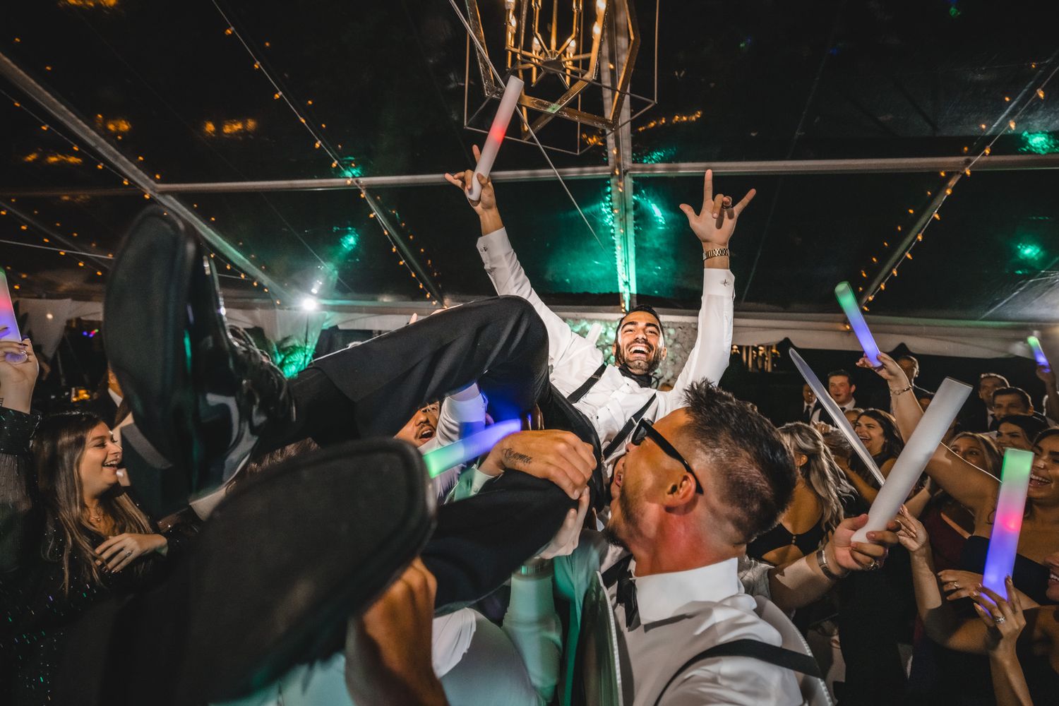 Concert crowd dances with raised arms under colorful stage lights during an energetic live music performance.