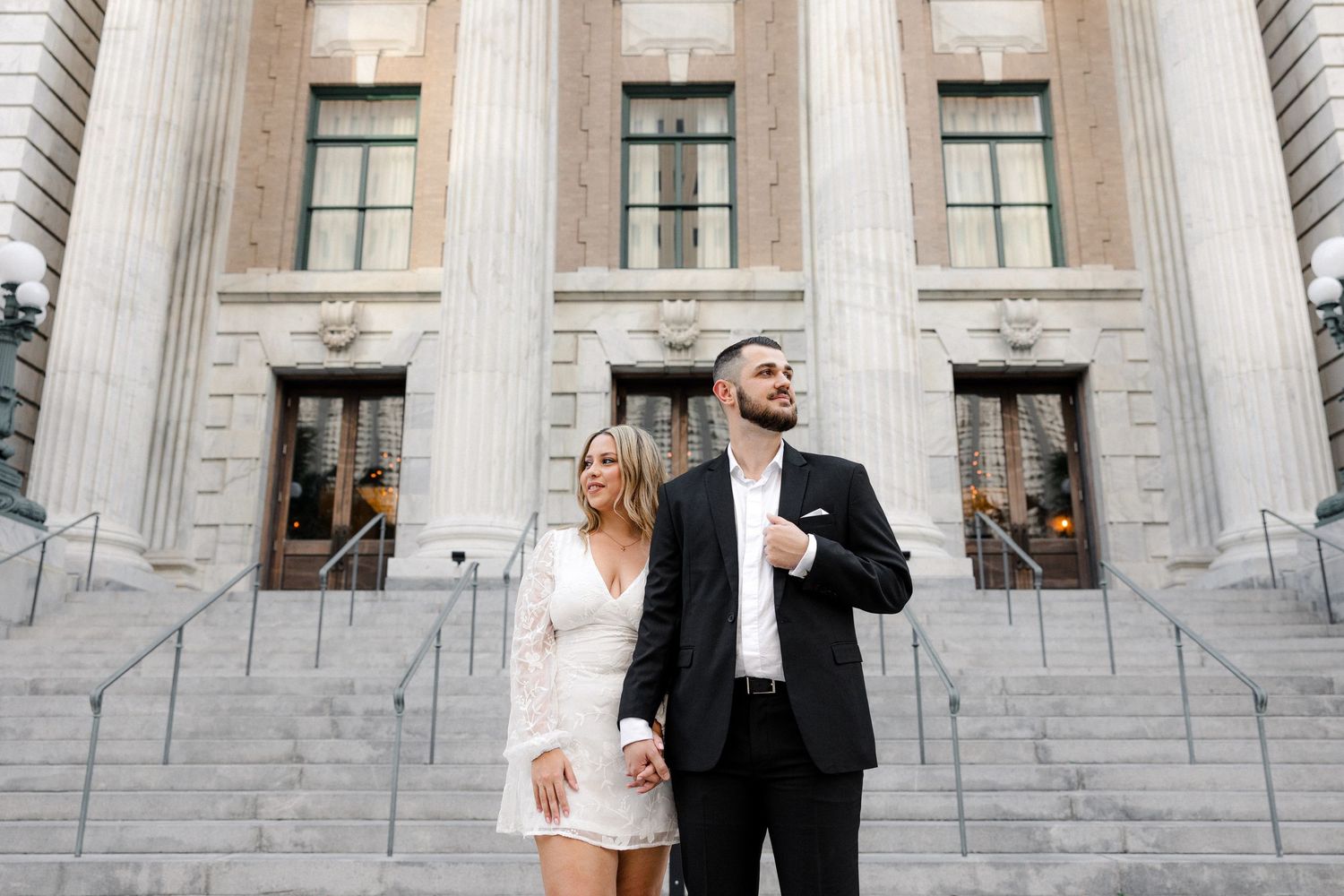 Couple in formal attire poses on grand building steps, woman in white mini dress, man in black suit holding hands.