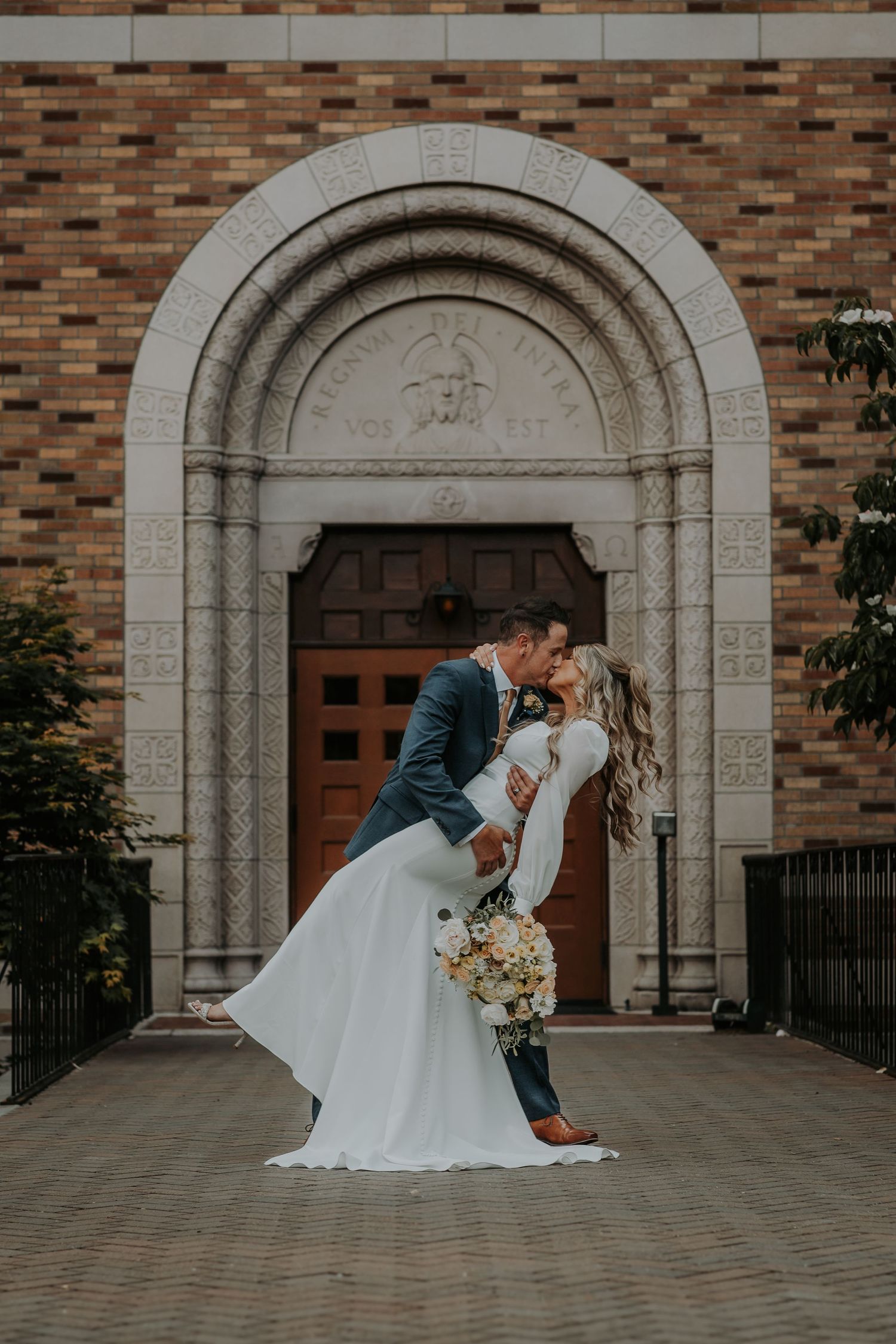 Groom in a navy suit dips his bride in a white gown holding a bouquet in front of a brick church with an arched wooden door.