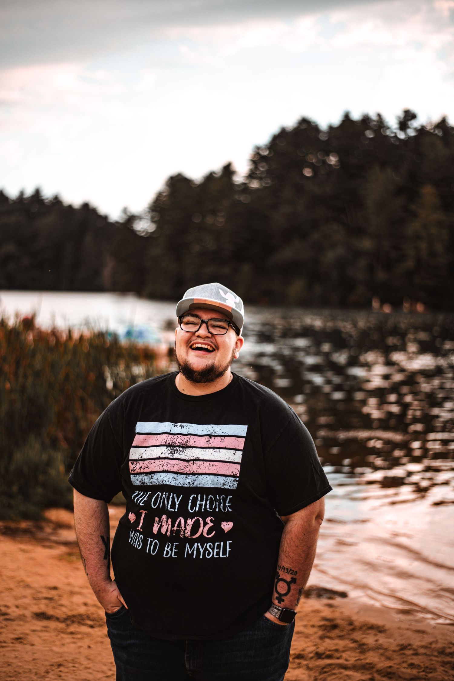 Person in glasses and graphic t-shirt smiling at sunset by a lake.