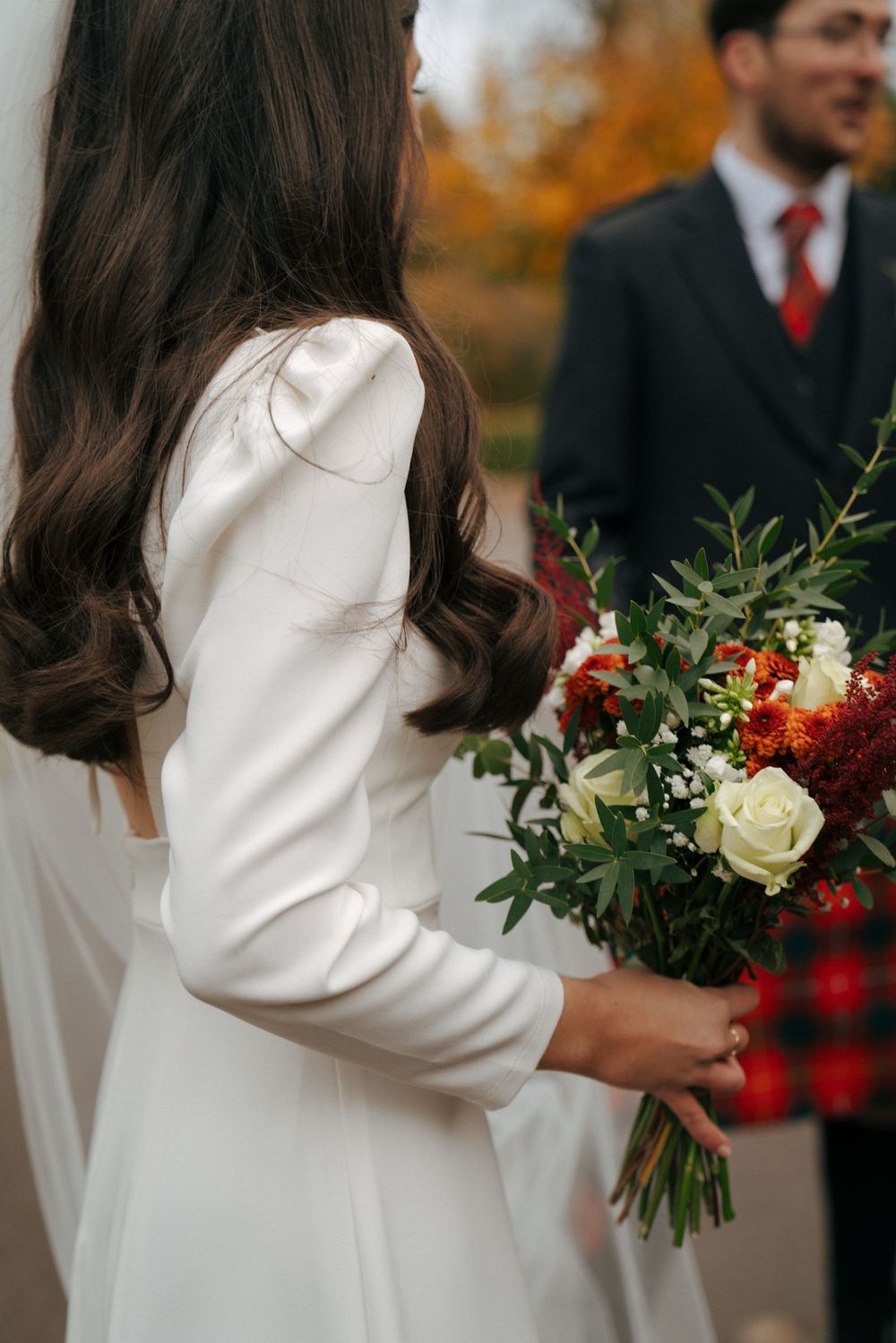Bride in long-sleeved white dress holding autumn-themed bouquet with red and white flowers.