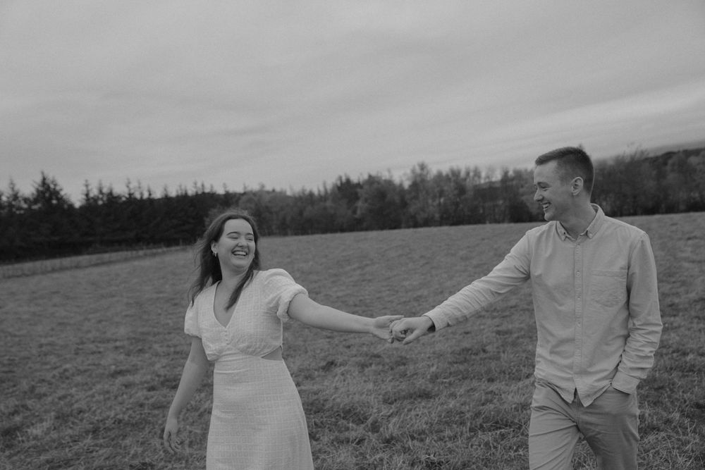 A couple in coordinated white and beige outfits dances in a scenic field during an outdoor photoshoot.