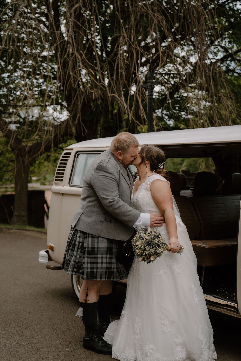 Couple shares intimate moment near vintage VW van under hanging tree branches.