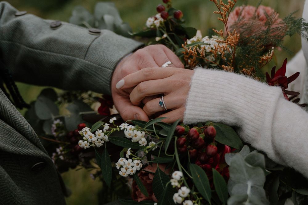 Couple holding hands with a blue sapphire engagement ring, surrounded by a lush bridal bouquet of red and white flowers.