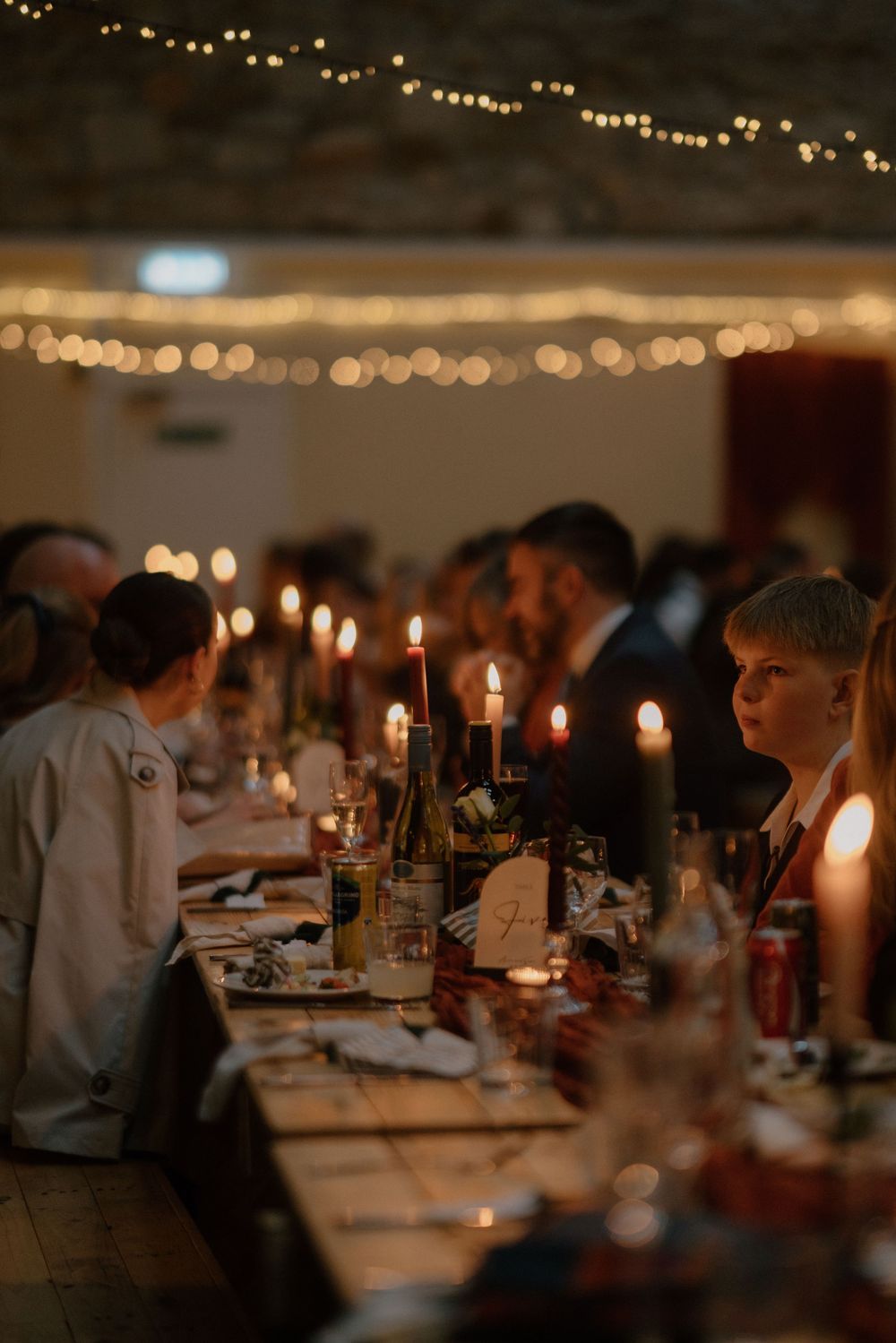 Guests seated at candlelit long tables during an elegant evening wedding reception with fairy lights overhead.