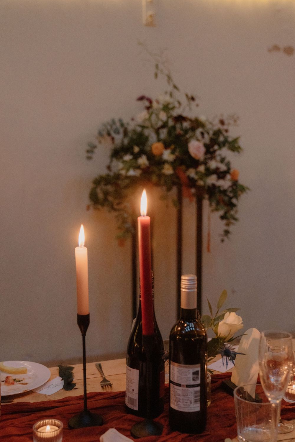 Romantic candlelit wedding table setting with wine bottles, lit taper candles, and floral arrangement in soft evening light.