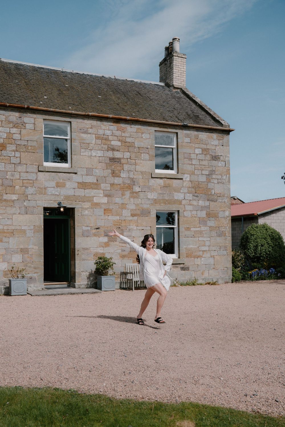 Woman in white dress dancing joyfully on gravel outside a traditional stone farmhouse under a blue sky.