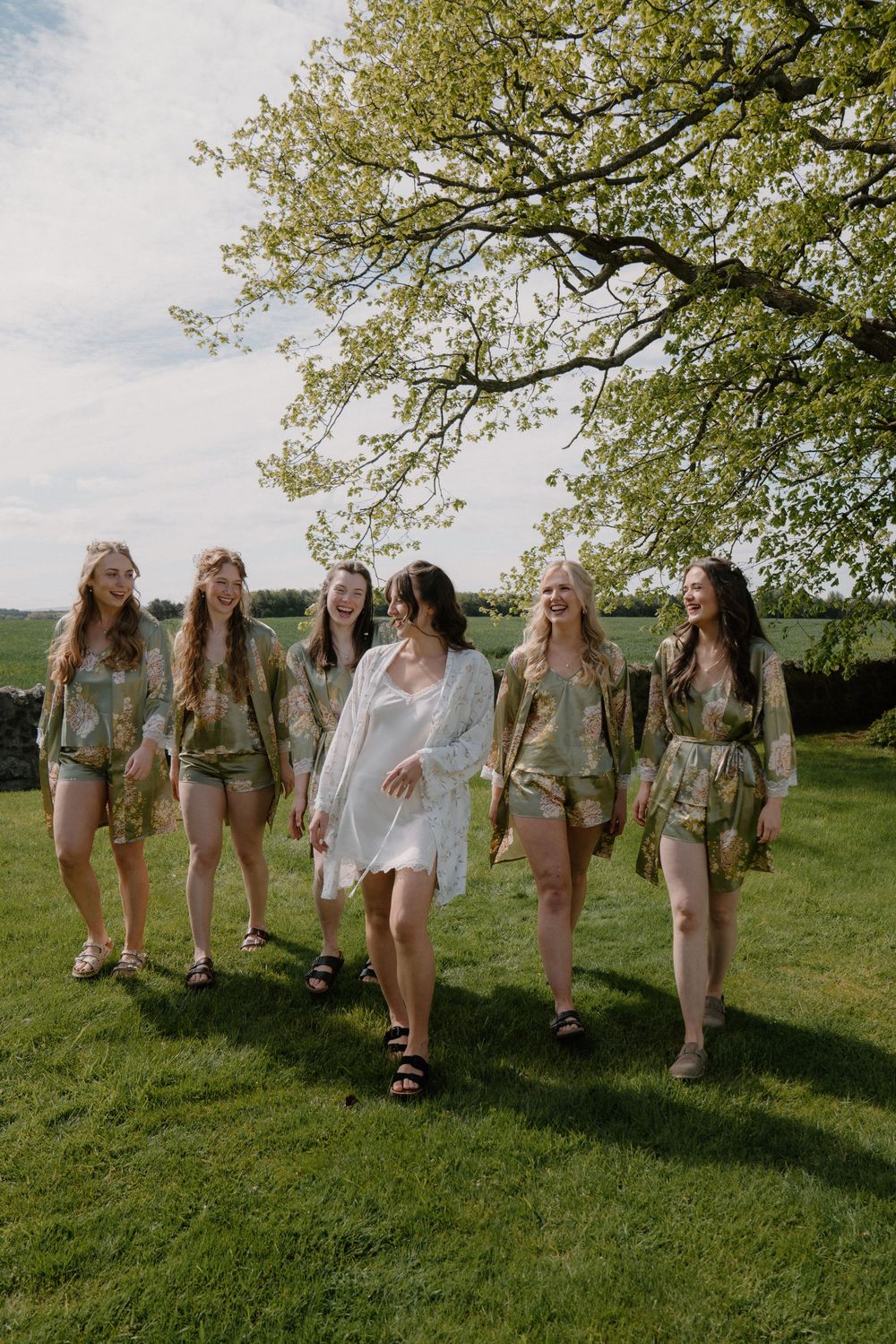 Bride in white robe walks with bridesmaids in matching green floral rompers on lush lawn under tree.