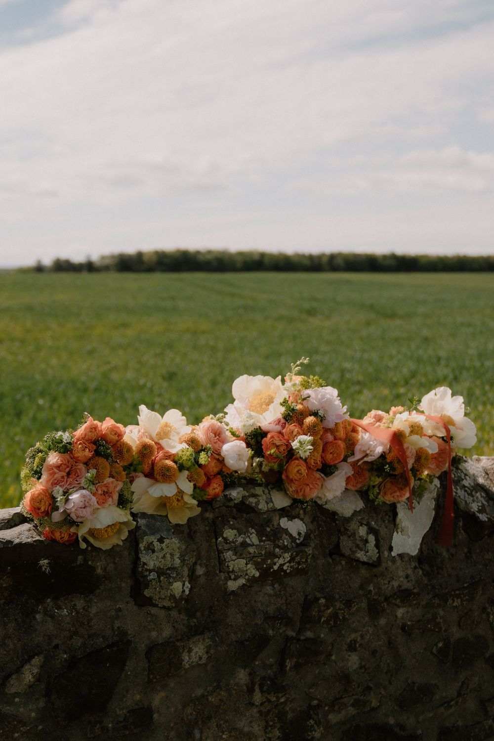 Peach and orange floral bouquets resting on a rustic stone wall with a lush green field and cloudy sky backdrop.