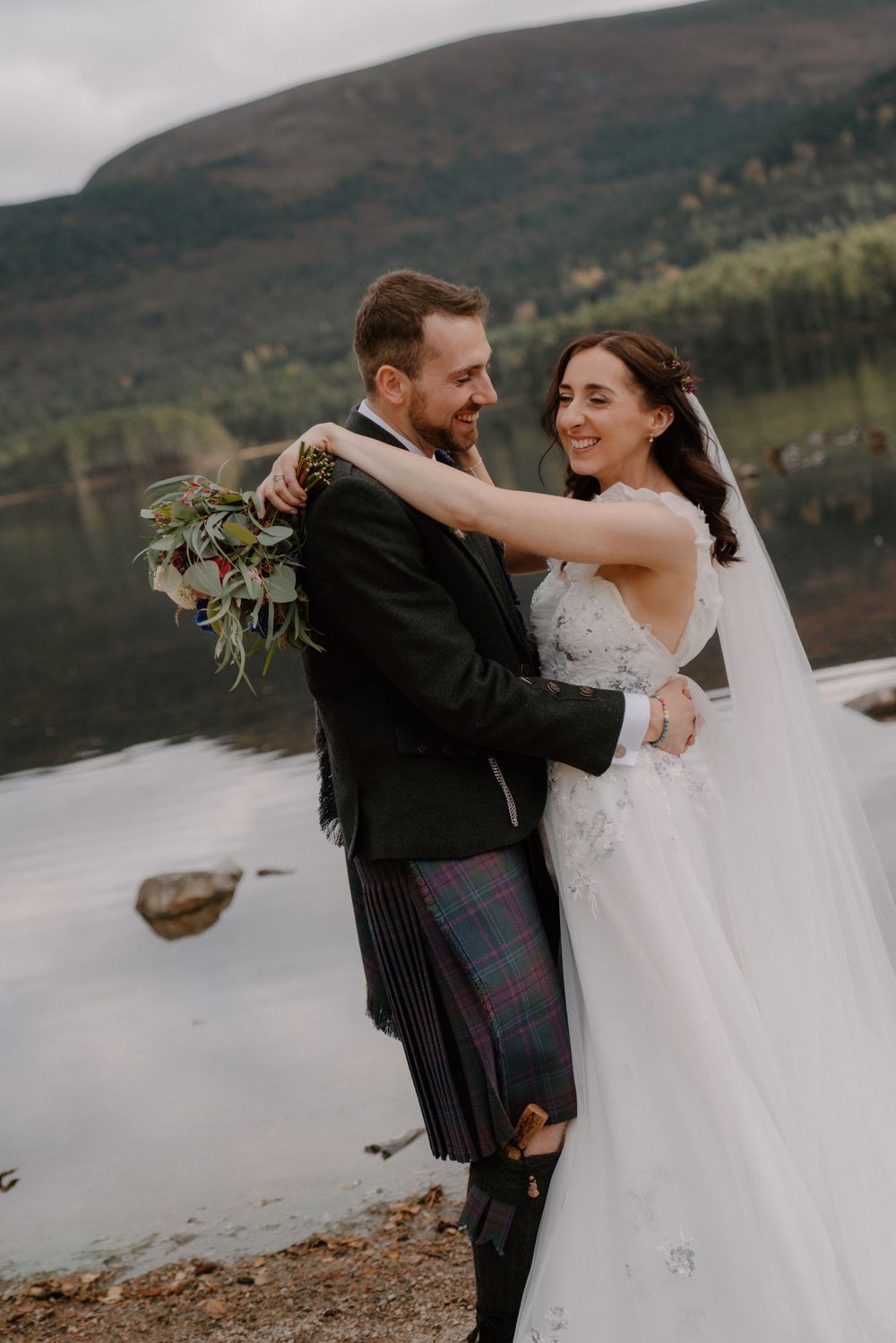 A couple embraces in wedding attire by a Scottish loch, groom in tartan kilt, bride in white gown holding bouquet.
