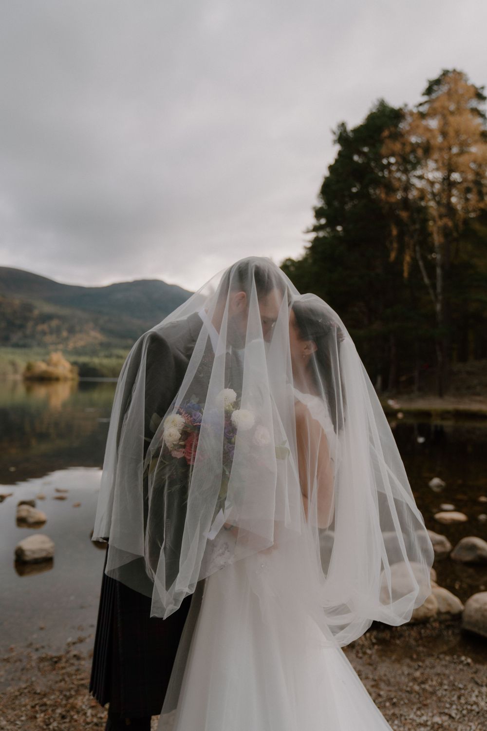A bride and groom share a kiss under a flowing wedding veil by a scenic lakeside with mountains in the background.